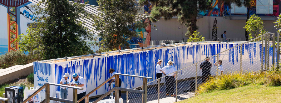 2 of 8, A series of large white canvases with blue painted lines on a walkway, with a colorful mural behind. 